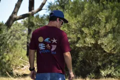 Remera de manga corta color bordó con un estampado gráfico en la espalda que incluye un sol amarillo, una ola azul, una estrella de mar blanca, un símbolo estilizado en azul y la frase "VITAMIN SEA" en azul.