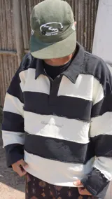 Buzo de tejido de punto grueso, estilo rugby, con cuello polo y tapeta de botones. Diseño a rayas horizontales anchas en blanco y azul marino, con bordes deshilachados en las uniones de las franjas. Presenta un logo bordado en el pecho.