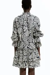 Vestido corto de algodón con estampado abstracto en blanco y negro. Tiene cuello con volados y lazo con borlas, mangas largas abullonadas con puños elásticos y falda escalonada con volados.