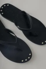 Sandalia plana tipo flip flop de cuero negro, con diseño minimalista y tira central de cuero. Presenta detalles de tachas metálicas en la base.