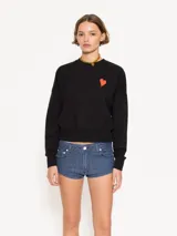 Buzo negro de algodón con cuello redondo y mangas largas. Presenta un pequeño corazón rojo estampado en el frente y un diseño de ángel en la espalda. Corte amplio y largo hasta la primera cadera.