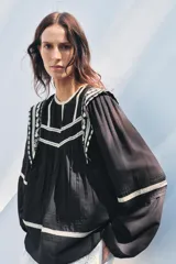 Blusa negra de algodón con cuello redondo y manga larga abullonada. Presenta bordados blancos a contraste en el cuello, hombros y mangas. Cierre en la espalda con abertura y botón.