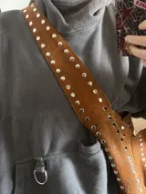 Cartera de cuero color caramelo con remaches metálicos, diseñada para uso cruzado o al hombro, con múltiples compartimentos.
