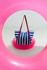 Bolso tipo tote bag de lona con rayas verticales azules y blancas. Posee asas largas de color rojo oscuro.