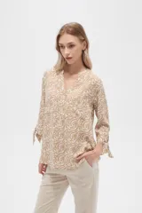Blusa color beige con estampado de arabescos blancos, cuello mao, mangas 3/4 con lazos en los puños, cierre parcial con botones y bolsillos frontales.