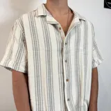 Camisa de manga corta, cuello cubano, color crudo con rayas verticales finas en gris oscuro. Presenta un bolsillo de parche en el pecho y botones marrones.