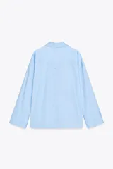 Camisa celeste de popelín con cuello solapa, escote pico y manga larga. Tiene bajo asimétrico con aberturas laterales y cierre frontal con botones.