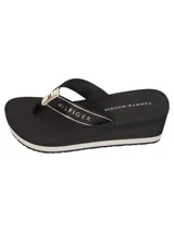 Ojotas de mujer con plataforma tipo cuña de color negro. Las tiras son de textil negro con una cinta decorativa que lleva el texto "HILFIGER" y el logo de Tommy Hilfiger en blanco y dorado. La plantilla lleva el logo de Tommy Hilfiger impreso.