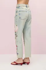 Pantalón de jean celeste con lavado vintage y calce mom. Presenta parches bordados con motivos florales, de mariposas y colibríes. El ruedo está desflecado.