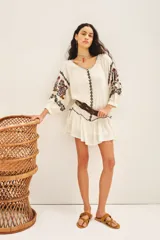 Vestido corto de bambula color blanco roto, con escote redondo y cartera frontal. Presenta bordados florales en tonos morados y marrones en el frente y mangas, y un volante en el ruedo. Se ajusta con un cinturón de tela a tono.