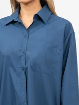 Vestido camisero corto, color azul, con cuello solapa, manga larga y bolsillo delantero de plastrón. Presenta un bajo asimétrico.