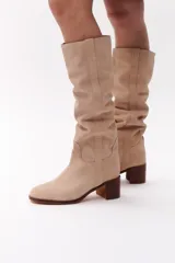 Bota de caña alta color beige, confeccionada en cuero gamuzado. Tiene taco de madera y puntera redondeada.
