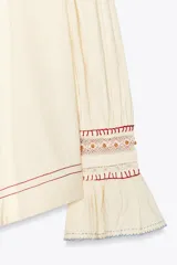 Camisa color crudo de cuello redondo con escote pico y lazada. Manga larga acabada en volantes. Detalle de bordados combinados a contraste y puntilla a tono.