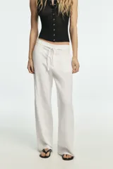 Pantalón blanco de tiro medio con pierna ancha y tejido que contiene lino. Presenta cintura con trabillas y un cinturón ajustable con lazada del mismo tejido. Cierre frontal con cremallera y botón.