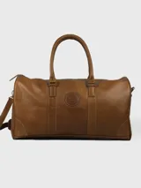 Bolso de viaje tipo duffel color marrón cuero, con doble asa corta y correa larga desmontable. Presenta un logo circular grabado en el centro y cierre superior con cremallera.