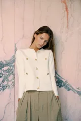 Chaqueta color crema de tejido de punto liso, con cuello redondo, manga larga, bolsillos delanteros de plastrón y cierre frontal con botones dorados.