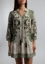 Vestido corto color crema con estampado de motivos geométricos en verde. Tiene cuello mao con lazo y borlas, mangas 3/4 y falda con volantes.