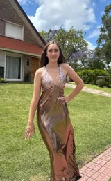 Vestido largo color marrón cobrizo con escote en V, cut outs laterales bajo el busto y breteles finos que se cruzan y atan en la espalda. Presenta un top con estampado abstracto en tonos marrones y dorados.