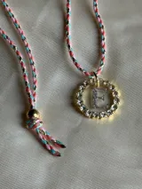 Collar con dije circular dorado con strass y letra 'A' en el centro, con cordón trenzado multicolor y cierre ajustable.