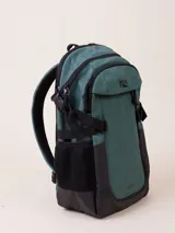 Mochila de nylon ripstop color verde bosque con detalles en negro. Presenta un compartimento principal con cierre, un bolsillo frontal con solapa y un bolsillo lateral de malla negra. Incluye correas ajustables en los laterales y un logo negro en la solapa frontal.
