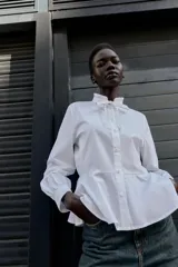 Camisa blanca de popelín con cuello redondo con volante y lazo, manga larga y bajo peplum. Cierre frontal con botones.