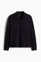 Camisa negra de tejido suave con cuello abierto, tapeta lisa y canesú en la espalda. Modelo de manga larga con cierre ajustable de botones en los puños y tapeta con botón. Bajo recto y corte estándar.