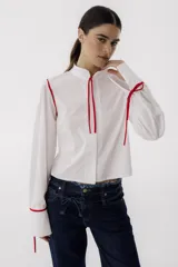 Camisa blanca de corte cropped con cuello mao y mangas largas abullonadas con lazos rojos en los puños. Presenta detalles de ribetes rojos en los hombros y lazos rojos en el cuello.