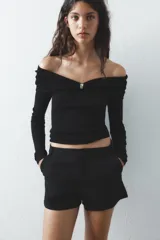 Top de punto negro con escote recto, hombros descubiertos, mangas largas y detalle de pieza metálica plateada en el delantero.