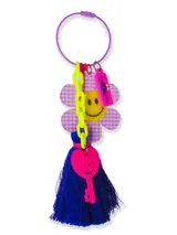 Llavero con aro metálico violeta y dijes colgantes: flor a cuadros lila y blanco con carita sonriente amarilla, cadena de eslabones verde flúor, etiqueta rosa con texto y llave rosa con carita sonriente. Incluye borla de hilos azul eléctrico.