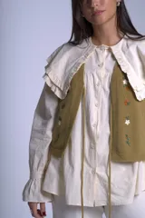 Chaleco tejido color crema con escote en V y cierre frontal con lazos. Presenta bordados de flores en tonos naranja, verde y blanco.