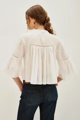Camisa blanca de algodón con cuello alto y volados, mangas largas abullonadas con puños elásticos. Presenta detalles de encaje y frunces en el frente y la espalda.