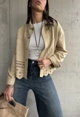 Sobrecamisa beige de gamuza con cuello camisero, cierre frontal con botones y mangas largas con puños abotonados. Presenta bolsillos de parche en el pecho y detalle de calado en el ruedo.
