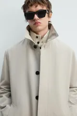 Gabardina color arena de corte recto, con cuello solapa, cierre frontal con botones, mangas largas con puños abotonados, bolsillos laterales y abertura en la parte posterior.