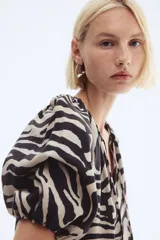 Blusa oversized de tejido liviano con estampado de cebra en tonos beige y negro. Tiene escote en V con cordón ajustable, mangas globo al codo de corte raglán y puños elásticos.