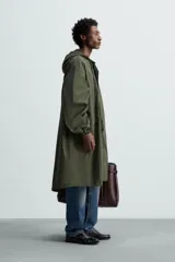 Parka verde militar de tejido técnico ligero, corte oversize, cuello alto con capucha ajustable, manga larga con puños elásticos, cintura ajustable con cordón interno, bolsillos con solapa, bajo asimétrico con abertura trasera ajustable y cierre frontal con cremallera oculta y botones a presión.