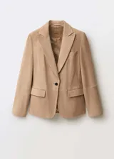 Blazer color beige de gamuza, con cuello de solapa, cierre de un botón, dos bolsillos frontales con solapa, mangas largas con puños abotonados y forro interior.