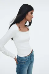 Remera blanca de manga larga, corte ajustado y corto (cropped), confeccionada en tejido acanalado. Presenta un escote pronunciado ribeteado en encaje y un pequeño moño de satén en el centro del escote.