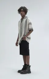 Camisa de manga corta color beige, tejida con punto abierto (símil crochet) y detalles en negro. Tiene cuello solapa, cierre frontal con botones y calce oversize.