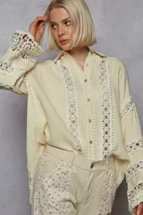 Camisa color crema de corte amplio, con cuello clásico, cierre frontal con botones y mangas largas tipo kimono con detalles de broderie en puños y frente.