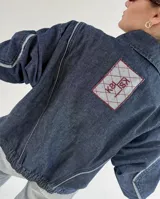 Campera de jean negra tipo bomber con cuello redondo y cierre frontal con botones metálicos. Presenta un parche rectangular con diseño gráfico en la espalda.