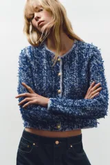 Chaqueta corta azul con tejido de punto entrelazado, cuello redondo, manga larga, bolsillos delanteros de plastrón y cierre frontal con botones dorados.