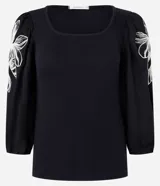 Blusa negra de rib con escote cuadrado, mangas largas abullonadas y bordado floral blanco en los laterales de las mangas.