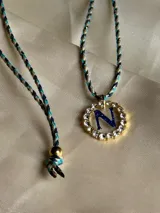 Collar con dije circular dorado con strass y letra A azul en el centro, colgando de un cordón trenzado multicolor.