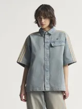 Camisa de manga corta Adidas Premium Essentials, confeccionada en denim azul claro 100% algodón. Diseño inspirado en la ropa de trabajo, con cuello clásico, cierre frontal con botones, bolsillo de parche en el pecho y logo bordado en el mismo tono. Presenta las tres tiras características de la marca en color beige en los hombros.
