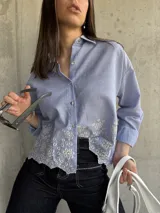 Camisa celeste a rayas verticales blancas, con cuello clásico, cierre frontal con botones y mangas largas. Presenta detalle de bordado calado con motivos florales en el ruedo.