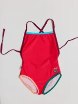 Traje de baño entero rojo con tirantes finos y espalda cruzada. Presenta un parche bordado con un monstruo en la parte inferior. Tiene ribetes en color borgoña y celeste en la parte superior, y ribetes en color naranja y verde en la parte inferior.