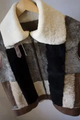 Chaleco sin mangas confeccionado con retazos de cuero en tonos marrón, beige, blanco y negro, con cuello de piel sintética color crema y cierre frontal con cremallera.