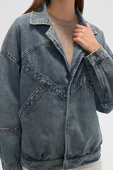 Campera de jean celeste con cuello clásico, solapas, cierre frontal con botones metálicos y mangas largas con puños abotonados. Presenta detalles de costuras diagonales y apliques de tachas metálicas.