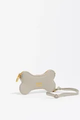 Monedero pequeño con forma de hueso, color gris claro, con cierre de cremallera dorada y correa de mano a juego. Presenta el logo de la marca estampado en dorado en el centro.