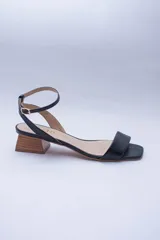 Sandalias de cuero negro con tira delgada sobre los dedos, pulsera al tobillo con hebilla dorada y taco geométrico color madera.